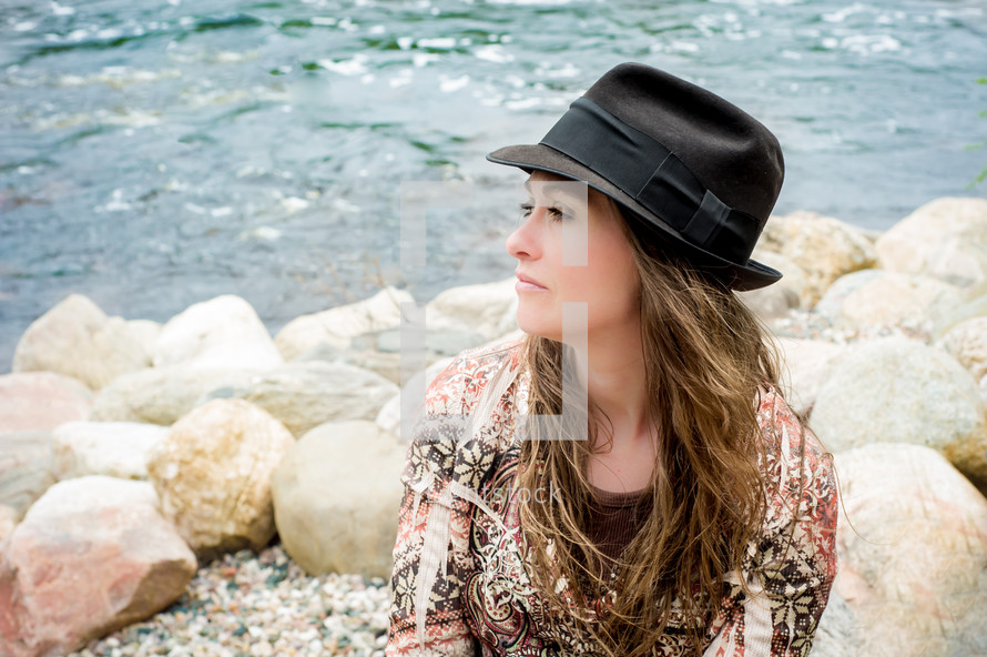 woman in a hat sitting at the rocky shore of a river