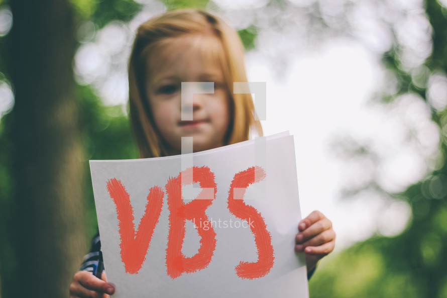 Child holding a handwritten sign saying "VBS."