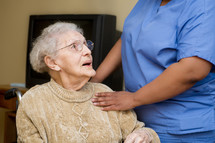 Hands of a caregiver touching the shoulder of an elderly woman in a wheelchair.