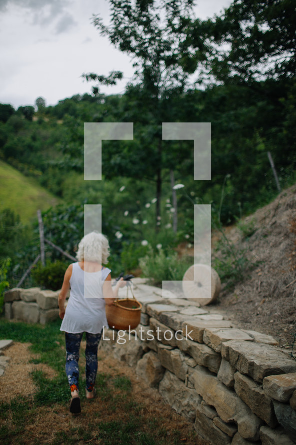 An older woman walking alongside a stone wall while carrying a basket.