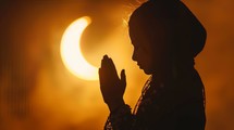 Silhouette of young Muslim girl praying with hands together against backdrop of crescent moon, symbolizing Ramadan, Islam, and spirituality.