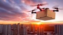 Quadcopter drone with cardboard box flying over the city at sunset