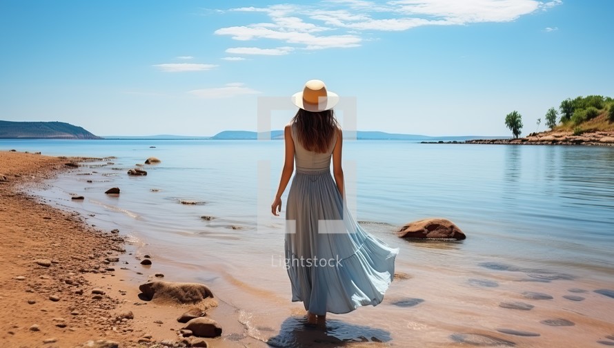 Beautiful girl in a white hat and blue dress walks along the shore of the beach