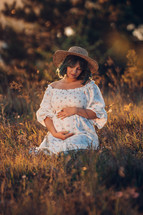 Happy Mother Carrying Child, Expecting For Baby. Amazing Motherhood. Pregnant Woman in beautiful dress and straw hat sitting on grass at nature rural meadow. High quality 