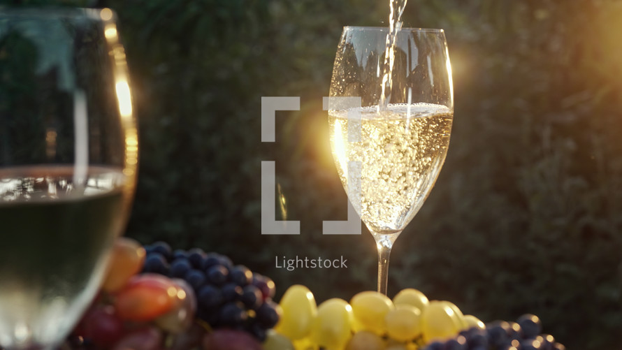 Stunning view of pouring white wine into glass goblet with fresh grape bunch background, sun light breaks through vineyard. High quality