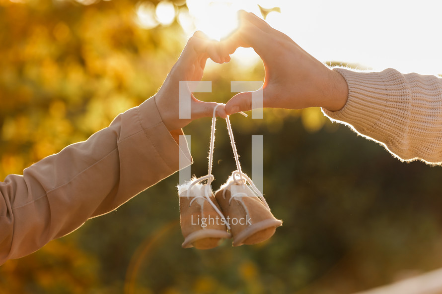 Man and pregnant wife are doing heart gesture with hands and holding warm baby shoes on nature autumn background. Future parents waiting for the baby. The concept of Mother's Day and Women's Day