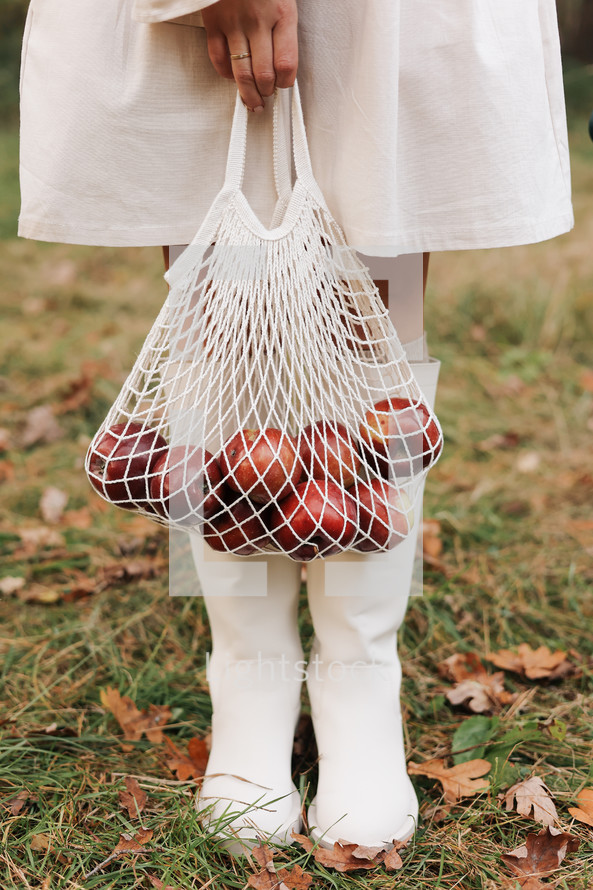 String bag with red apples. View of female legs in white boots walking through the autumn garden with eco bag in hands. Zero waste concept. No plastic life. Healthy fitness lifestyle. Recycling waste.