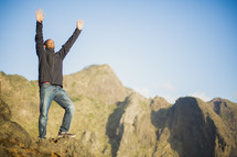 African American man standing on rock cliff with raised hands 