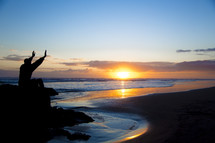 man sitting on a rock on a beach at sunset with raised hands 