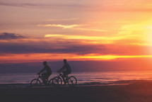 Two male cyclists bike down the beach at sunset. 