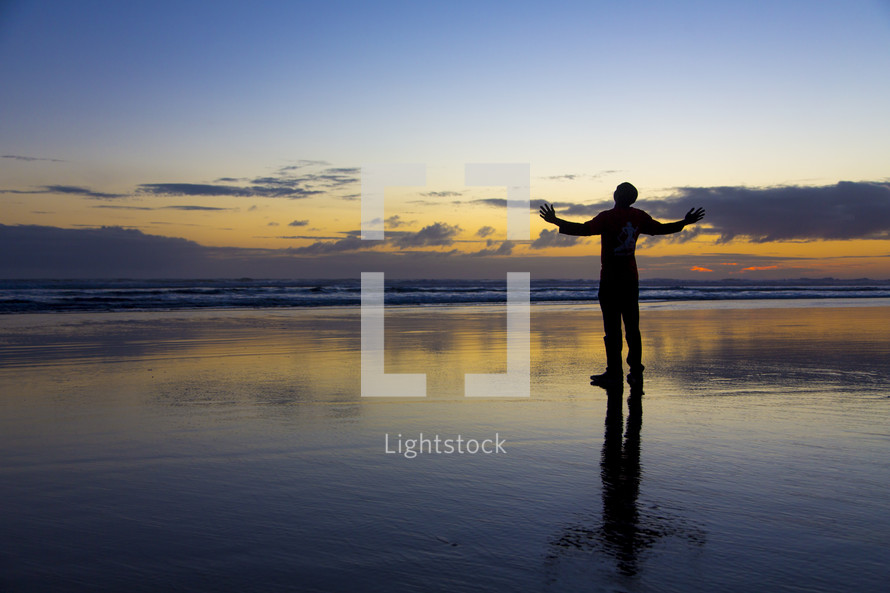 silhouette of a man standing on wet sand with hands raised in worship 