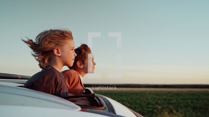 Adorable happy little kids stands in open car sunroof during road trip in countryside, rural landscape at summer. Concept of family leisure, active traveling. High quality