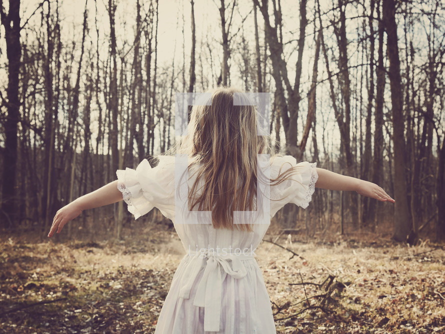 a girl in a dress standing with arms out-stretched in front of a forest 