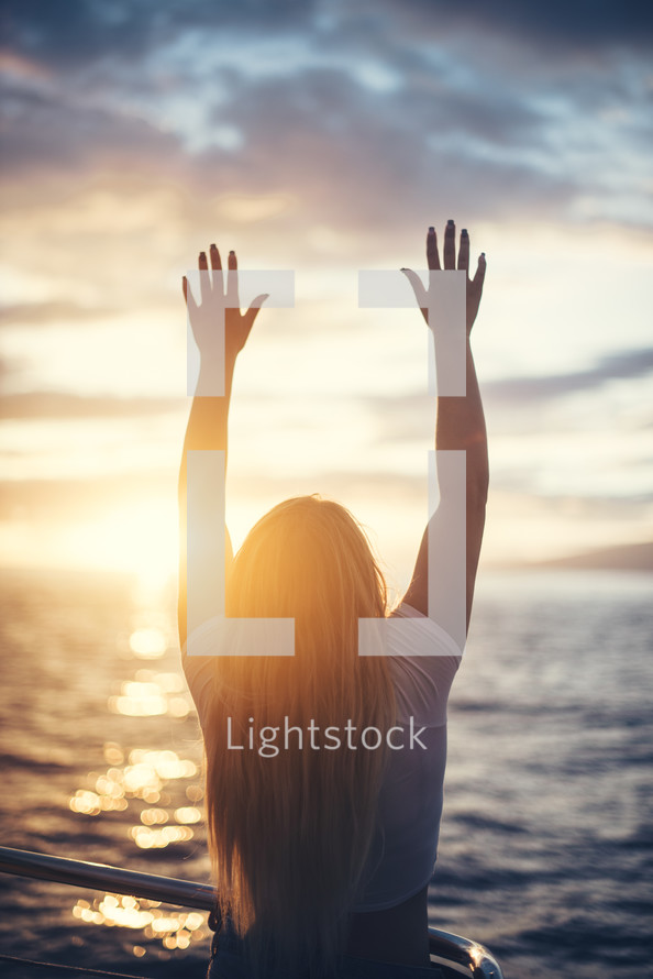 girl with raised hands standing in front of the ocean on a beach at sunset 