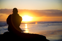 silhouette of a man sitting on a rock at a shore at sunset 
