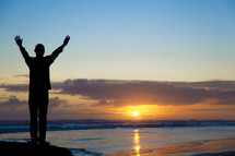 silhouette of man standing on a beach with hands raised in worship 