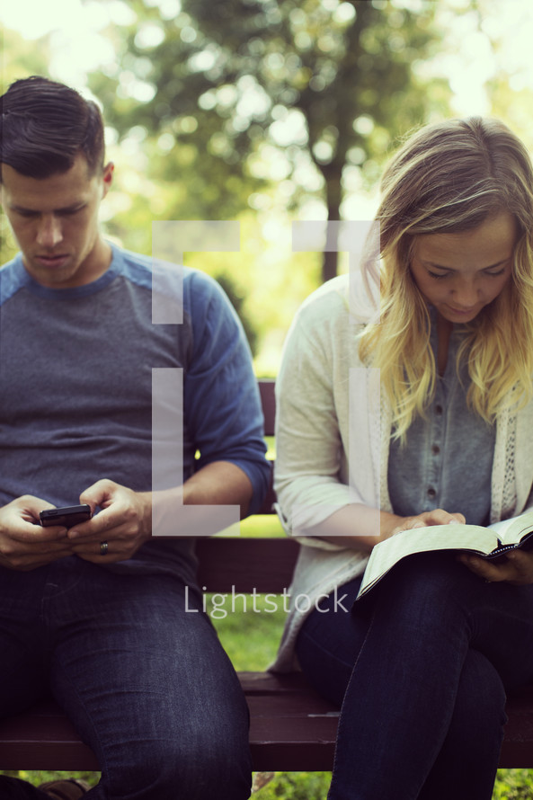 a couple sitting on a park bench, reading and texting 