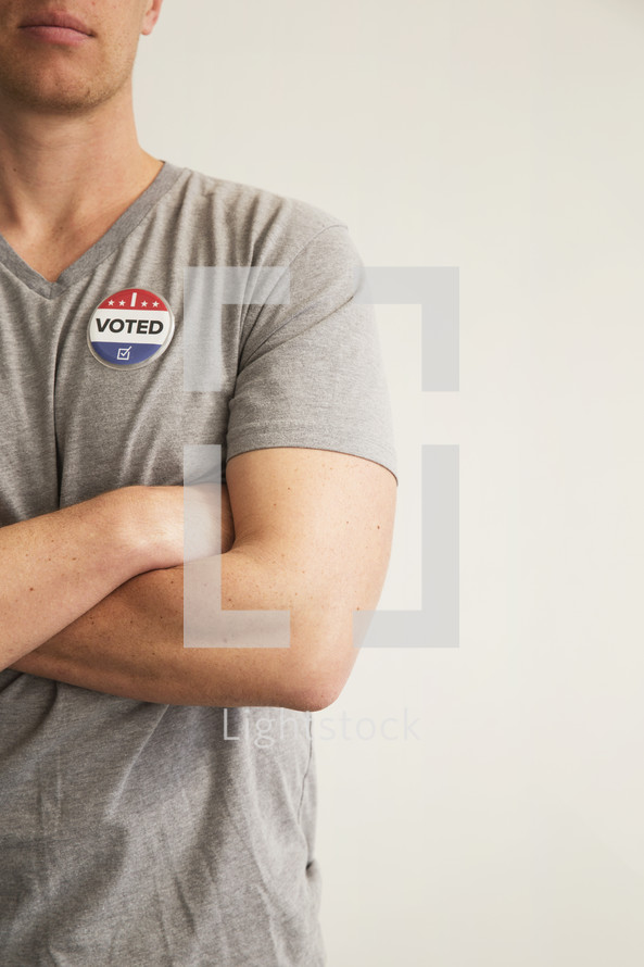 A man with arms crossed wearing a red white and blue button reading, "I Voted."