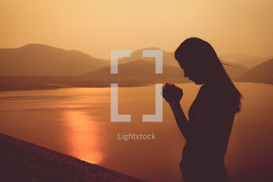 silhouette of a woman praying by a lake at sunset 