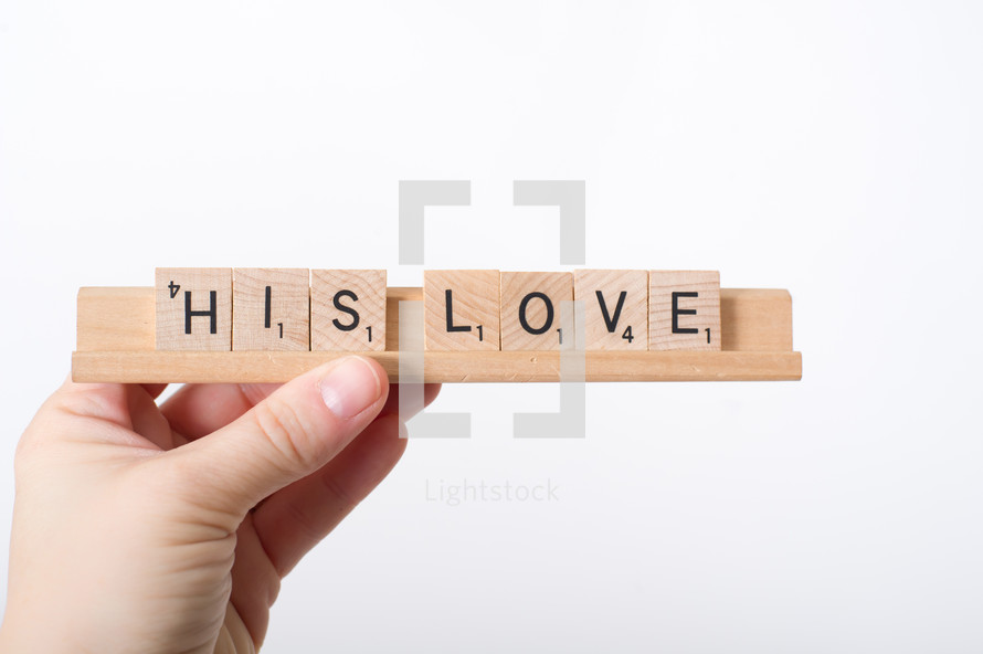 A hand holding scrabble pieces with the words his love. 