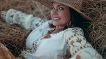 Pretty woman in straw hat and embroidered blouse smiling lying on hay in countryside at sunset. Rural nature, haystack, vacation, relax and harvest concept.