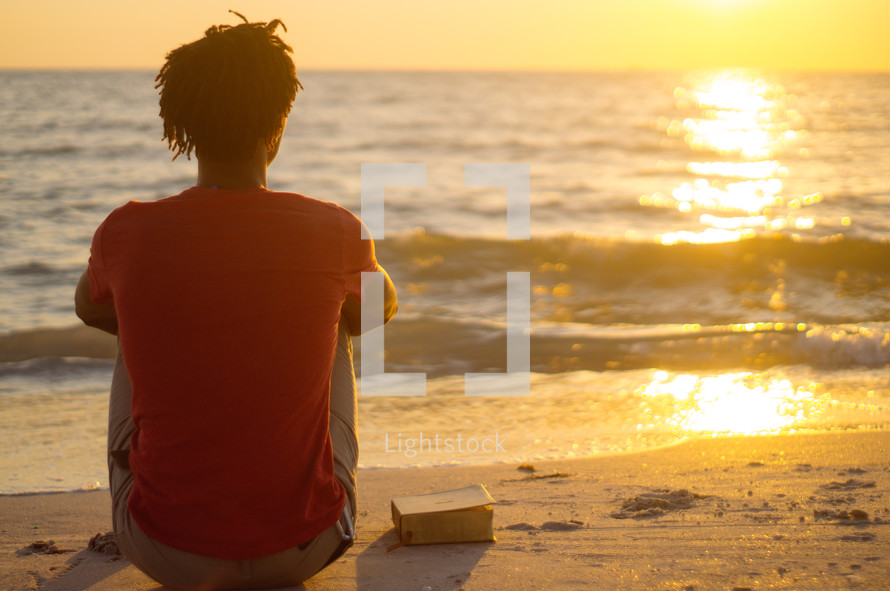 African-American man sitting on a beach with a Bible meditating 