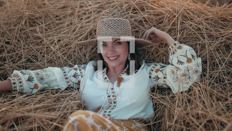 Pretty woman in straw hat and embroidered blouse smiling lying on hay in countryside at sunset. Rural