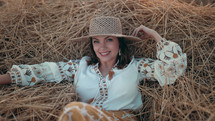 Pretty woman in straw hat and embroidered blouse smiling lying on hay in countryside at sunset. Rural