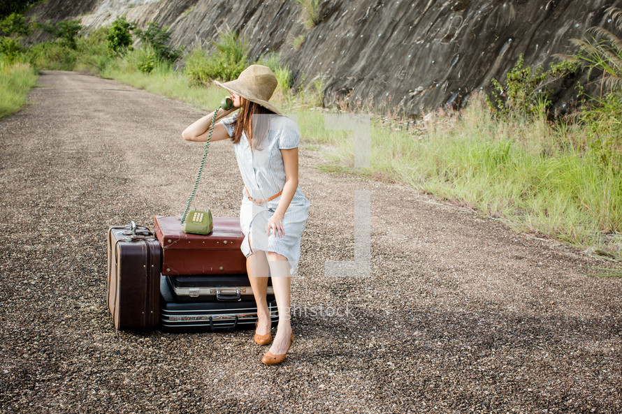 a woman sitting on a dirt road next to luggage and talking on a vintage phone 