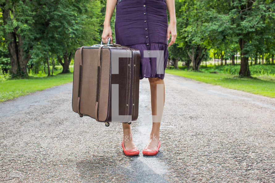 woman standing on a rural road holding luggage 