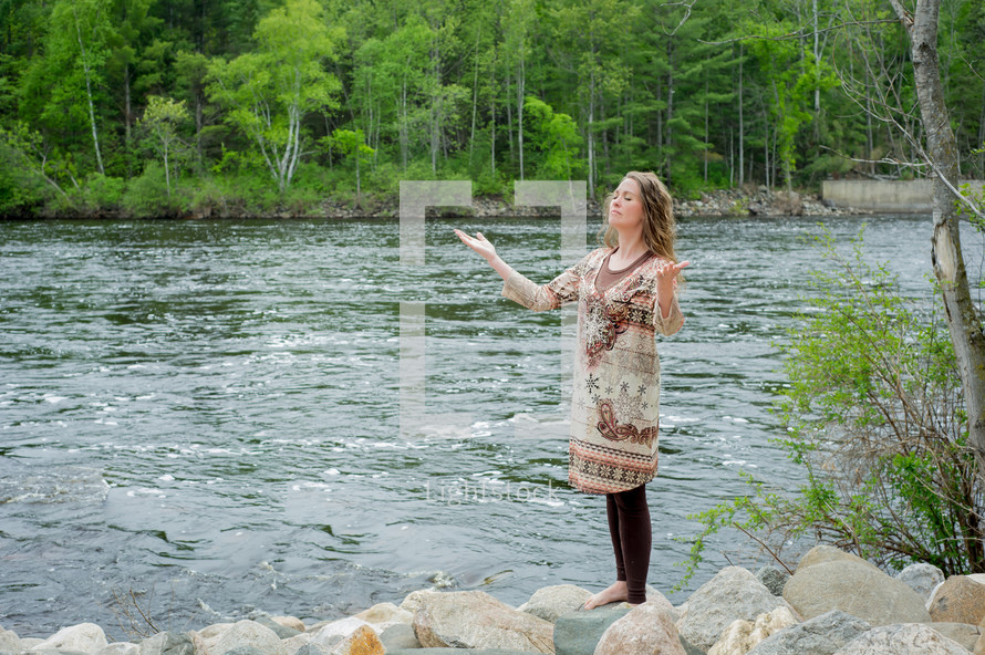 woman standing at the shore of a river with her hands raised in praise and worship to God