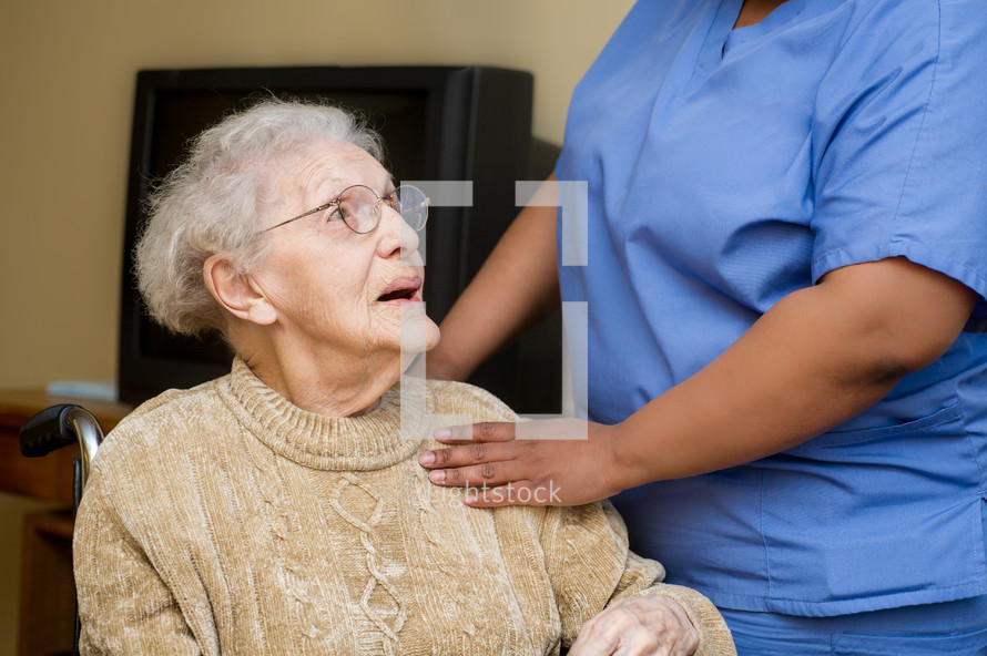 Hands of a caregiver touching the shoulder of an elderly woman in a wheelchair.
