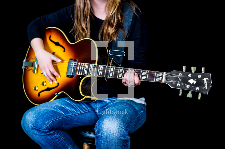 Woman sitting on stool playing a guitar.
