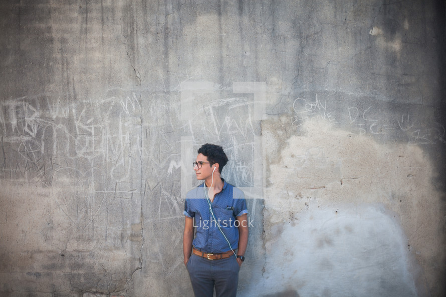 man listening to earbuds standing in front of a gray wall with graffiti 