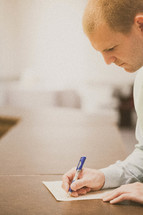 Man with pen in hand writing on a piece of paper on a desk.