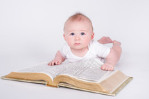 Baby dressed in white, laying on the floor with his propped on a giant bible