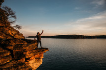 a man with a raised arm standing on the edge of a cliff at sunset 