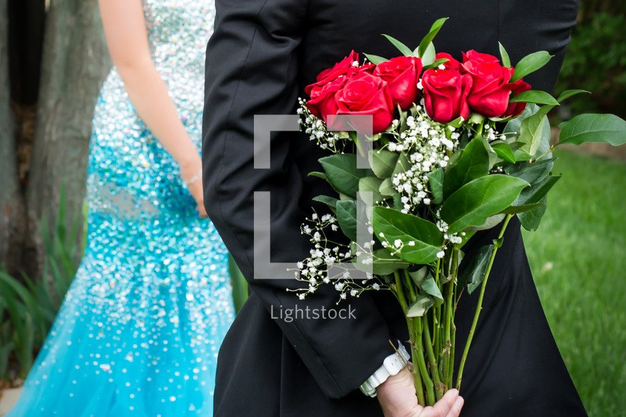 A man holding a bouquet of red roses behind his back for his date. 