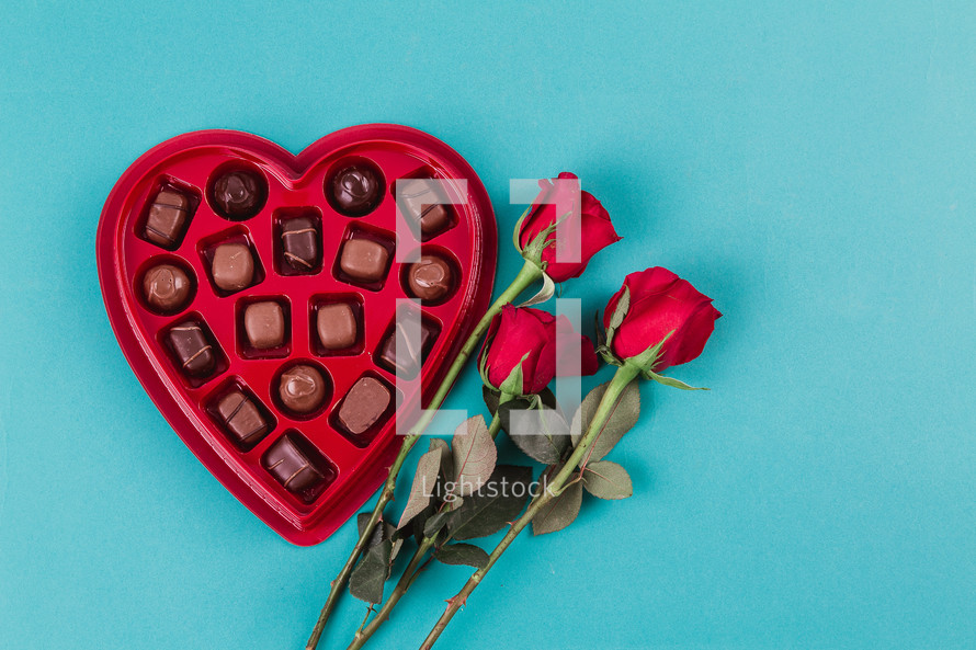 A heart shaped box of chocolates and three red roses.