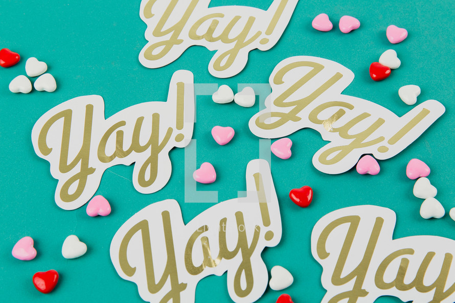 Small candy hearts and paper cutouts with the word "yay!" on an aqua background.