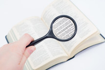 Hand holding a magnifying glass on a page of the Bible open to 1 Corinthians.