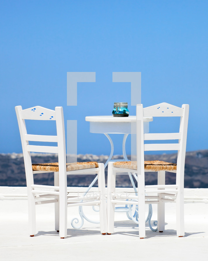 Two white chairs and a blurred panoramic view of the hills of santorini, Greece.