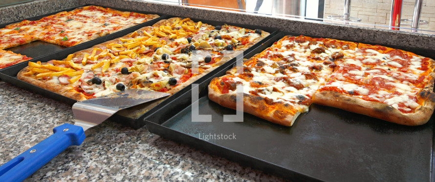 Counter of the pizzeria with trays and square pieces of pizza.