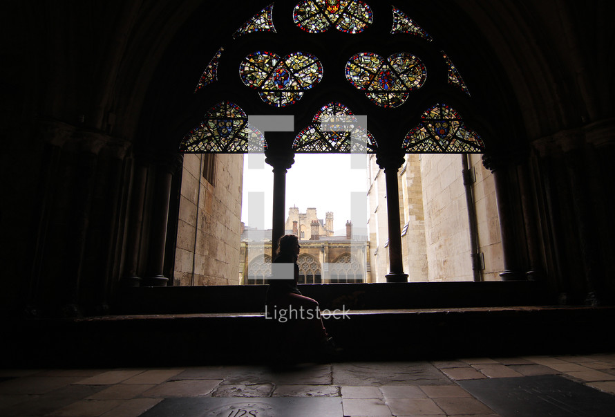 Silhouette of a woman sitting near a stained glass window in an ancient cathedral.