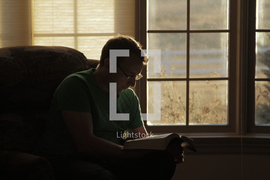 Man sitting in a chair in front of a window reading the Bible.