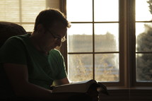 Man sitting in a chair reading his Bible by the window.
