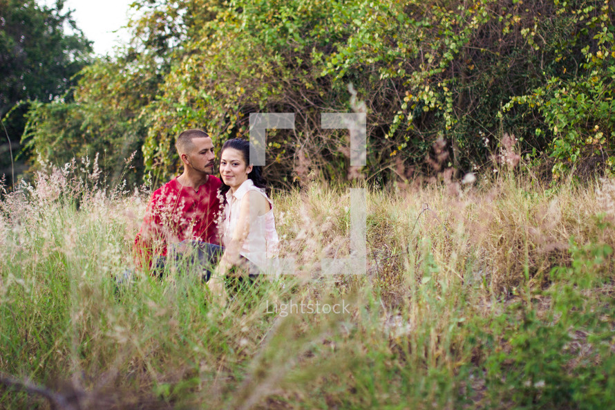 Couple sitting in a weedy field, trees in the background