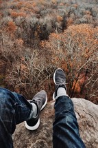 feet hanging off the side of a mountain over fall trees in a forest 