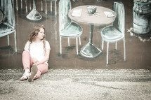 woman sitting on a sidewalk in front of a painting of table and chairs on a wall 