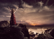 woman standing on a rock ledge near the ocean with her arms raised to the Lord in praise and worship